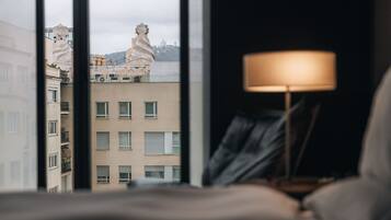 Una habitación con vistas a edificios a través de una ventana, una lámpara sobre una mesita de noche y una cama.