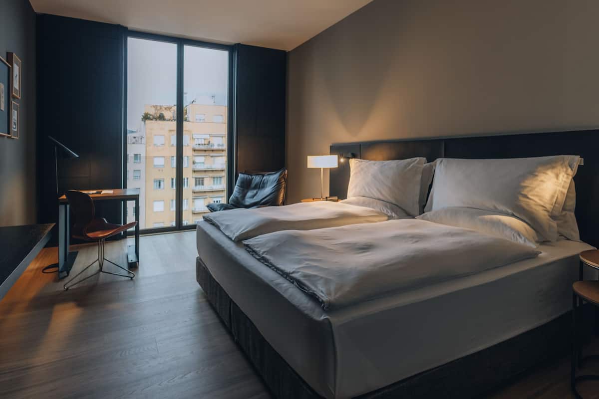 Habitación de hotel con una cama grande, un escritorio, una silla y vistas a edificios a través de un ventanal amplio.