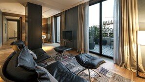 A modern living room with a sofa, ottoman, and a television. There is a large window with curtains, a door leading to another room, and a view of the outside. - Alma Barcelona GL (Barcelona)