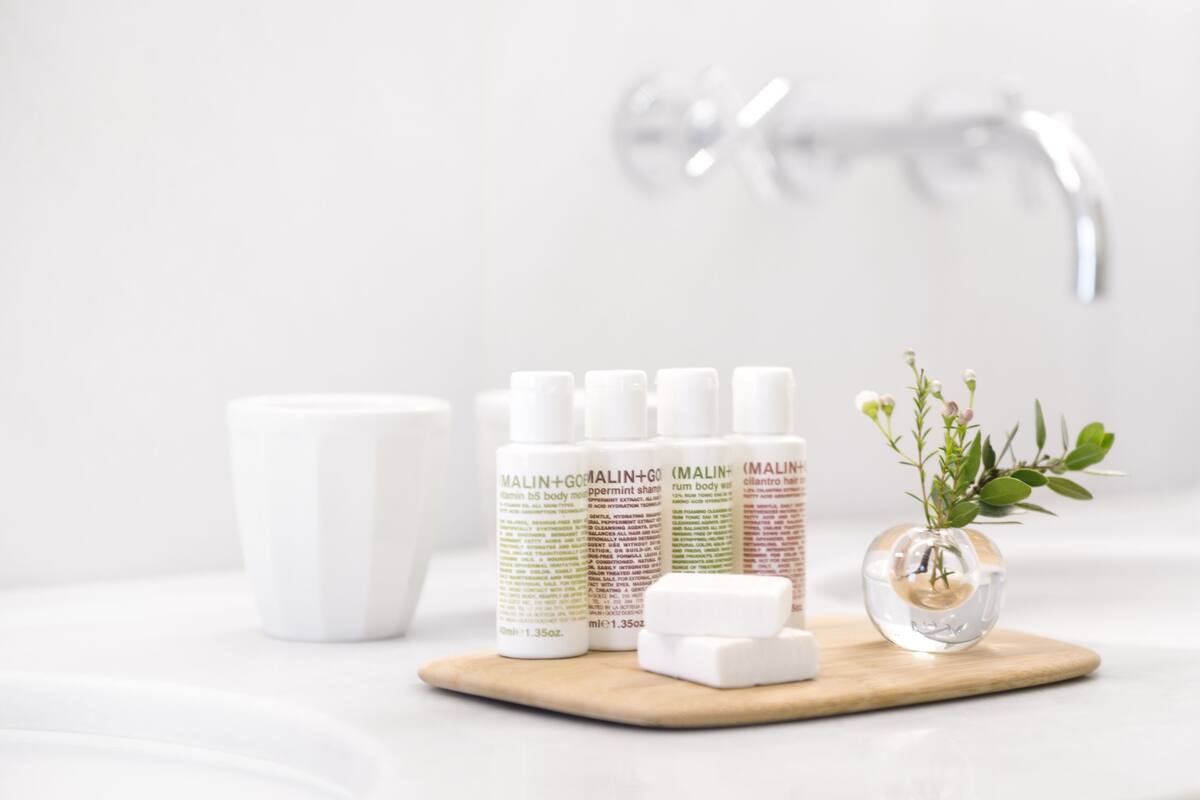 Un baño con lavamanos blanco, una bandeja de madera con productos de cuidado personal y un pequeño florero con flores.