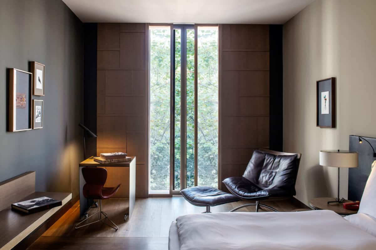 Una habitación de hotel moderna con un ventanal grande, un escritorio, un sillón de cuero, una cama y una mesita de noche.