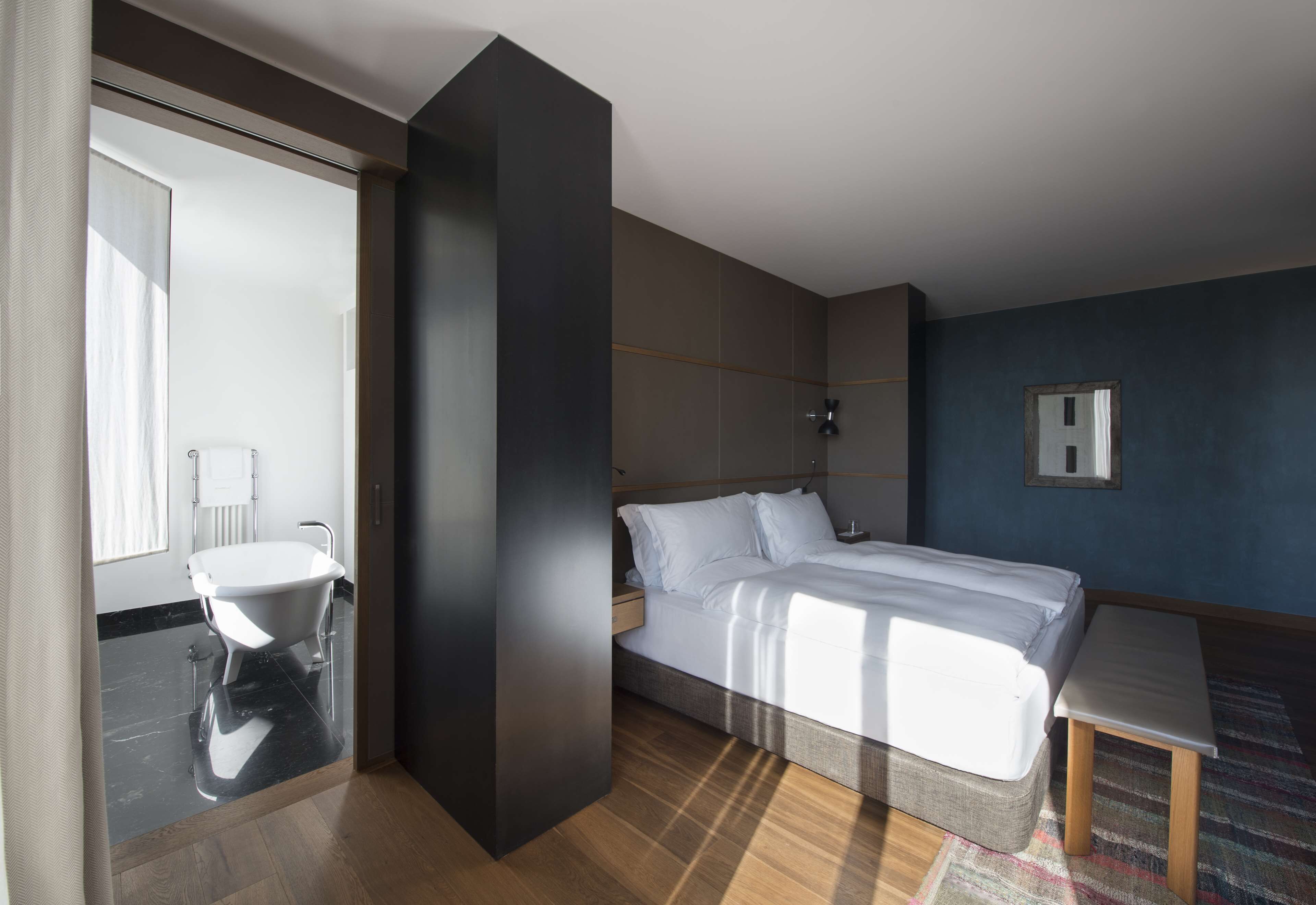A modern hotel room with a large bed, a bedside table, a small table, a window, and a bathroom with a bathtub.