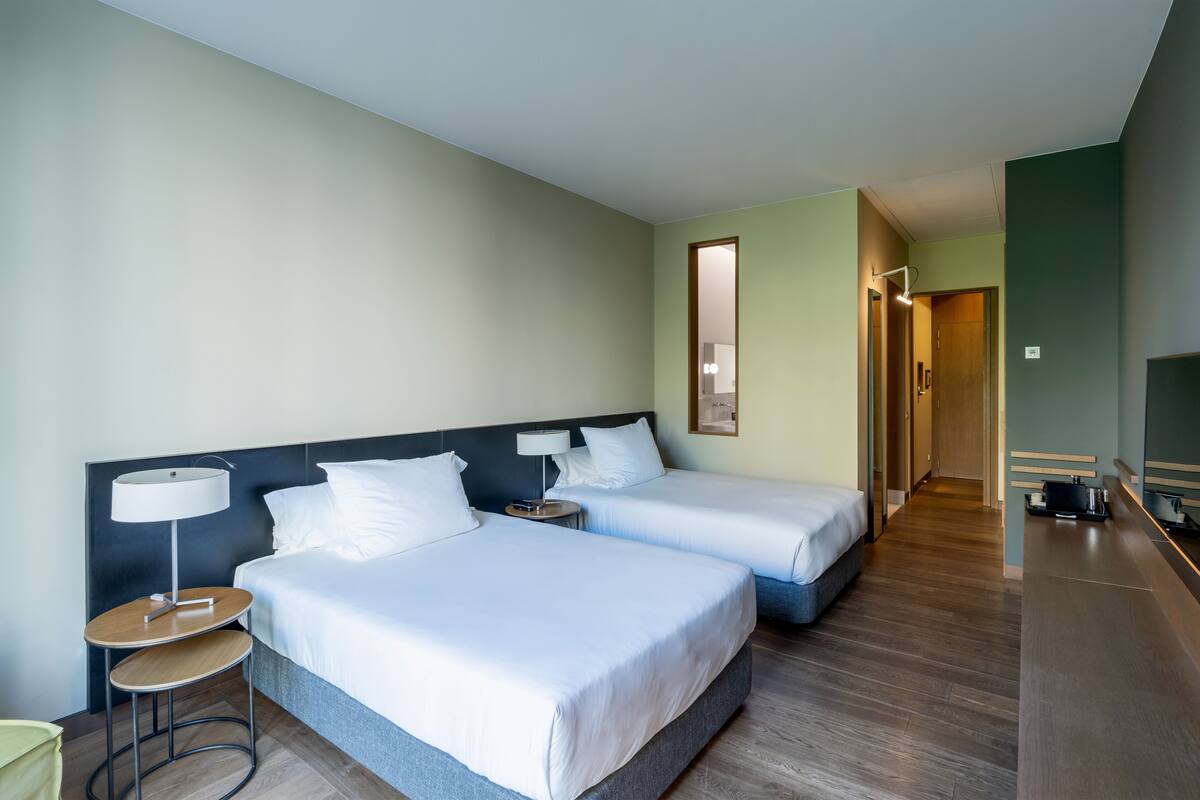 Una habitación de hotel con dos camas, una mesita de noche con lámpara, un taburete pequeño y un espejo.