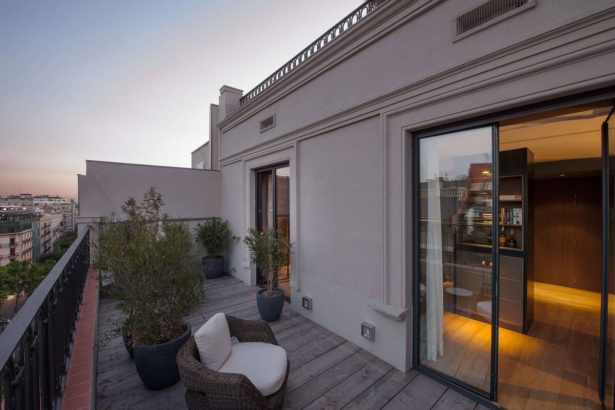 Un balcón con una silla de mimbre y plantas en macetas, una puerta de cristal que da a un espacio interior con zona de bar y vistas a la ciudad al fondo.