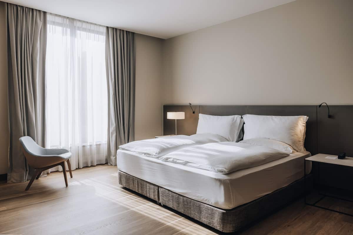 Una habitación de hotel moderna con una cama grande, una silla, una mesita de noche y una ventana con cortinas.