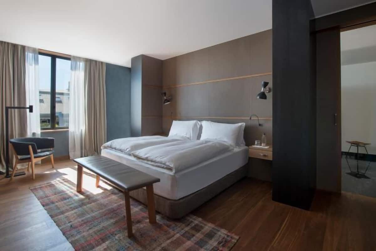 Habitación de hotel moderna con una cama grande, mesitas de noche, un banco, una silla y vistas a la ciudad.