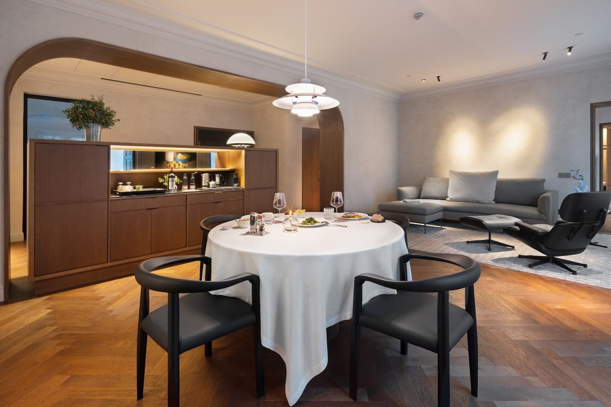 Un comedor moderno con una mesa redonda, sillas negras y un mantel blanco. Se observa una cocina con armarios de madera y una planta sobre la encimera.