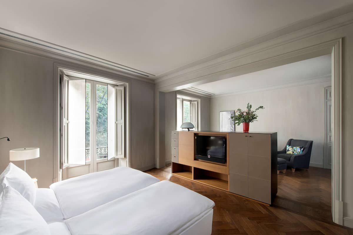 Habitación de hotel con una cama grande, un televisor sobre un mueble de madera, una silla con un cojín de colores y una planta en maceta.