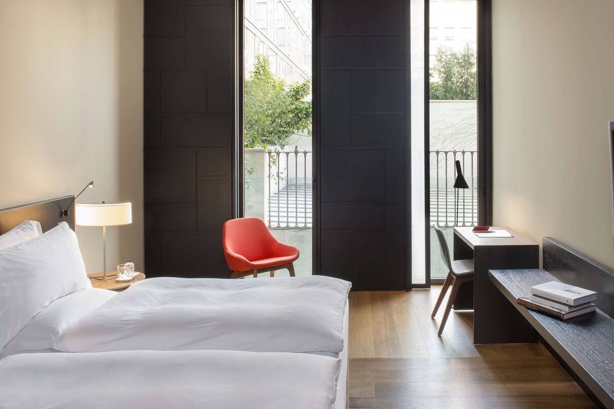 Una habitación de hotel moderna con cama, escritorio, silla, televisor y grandes ventanales con vistas a la ciudad.