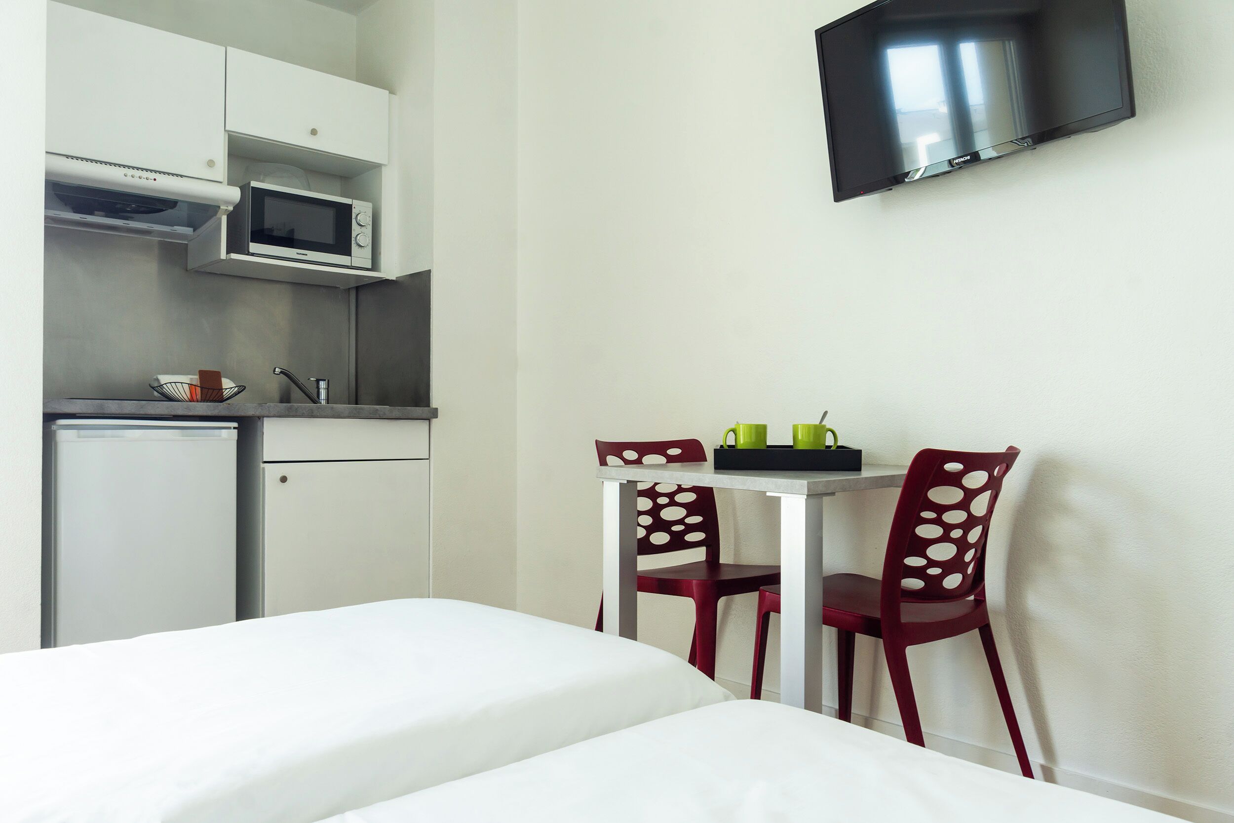 A compact room with a bed, a small kitchenette with a microwave, a TV mounted on the wall, and two chairs with a table.