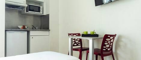 A compact room with a bed, a small kitchenette with a microwave, a TV mounted on the wall, and two chairs with a table.