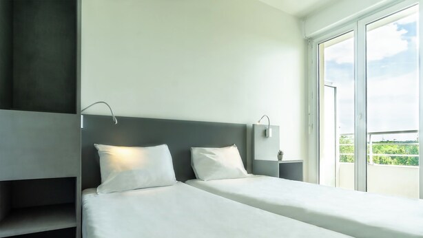 A modern hotel room with a large bed, a bedside table, and a window with a view of greenery. - City Residence Chelles (Chelles)