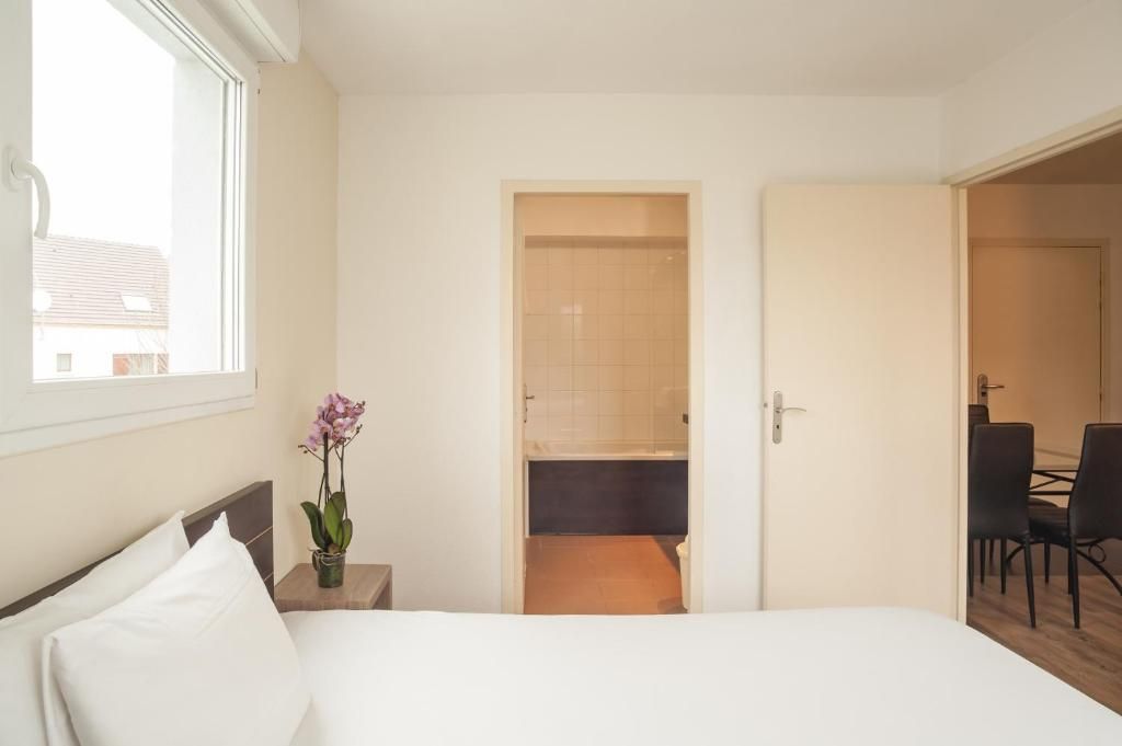 Una habitación de hotel con una cama, una mesita con un jarrón de flores, un baño con ducha e inodoro, y un rincón comedor con sillas.