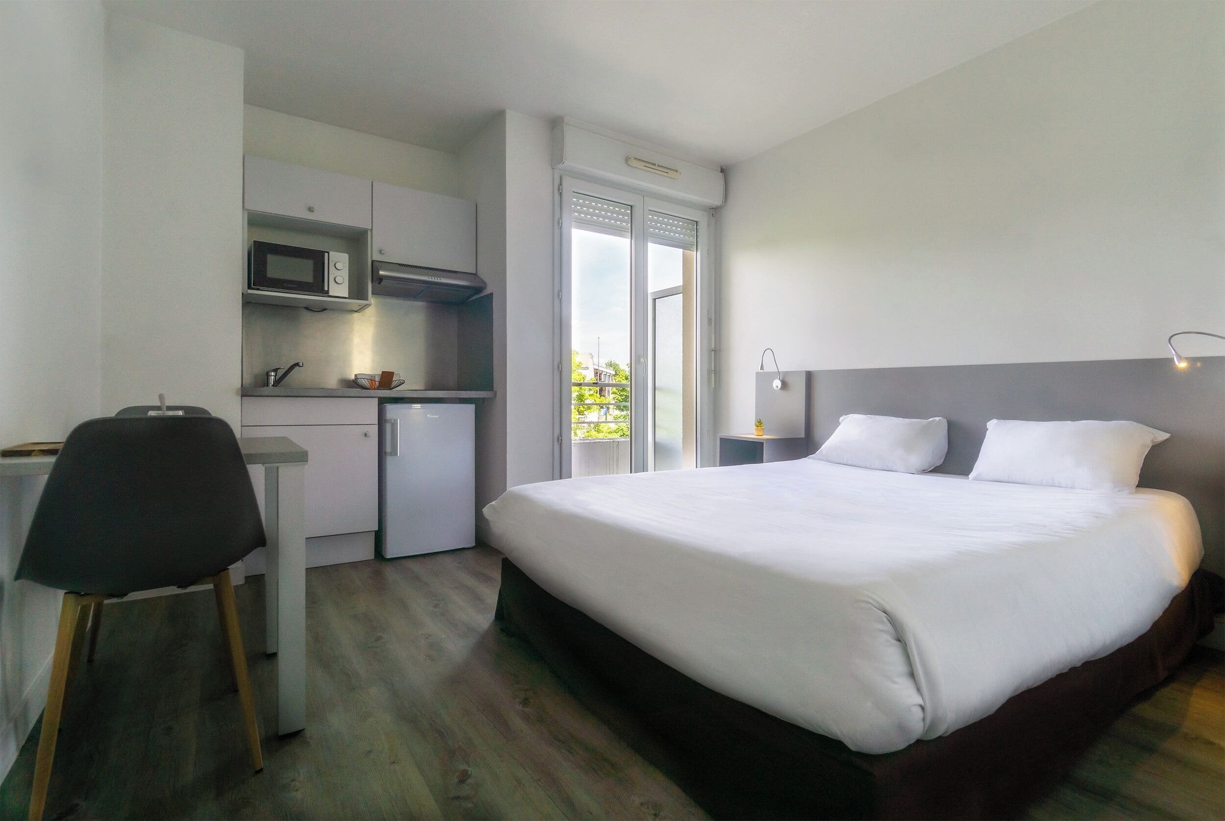 Une chambre dotée d’un grand lit, d’un bureau avec une chaise, d’une kitchenette et d’une fenêtre donnant sur un paysage verdoyant.