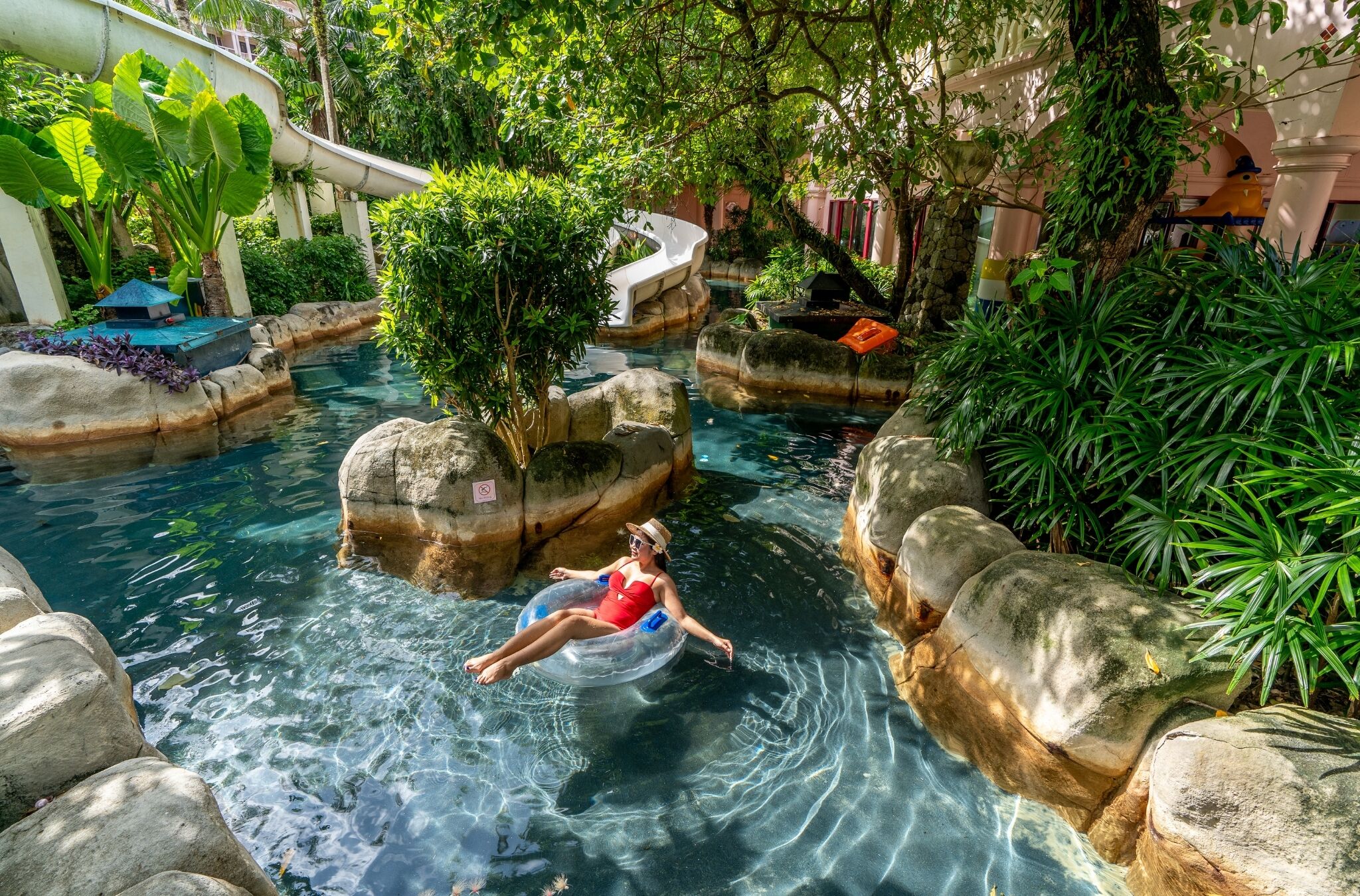 A pool area with a person floating on an inflatable ring, surrounded by rocks and greenery.