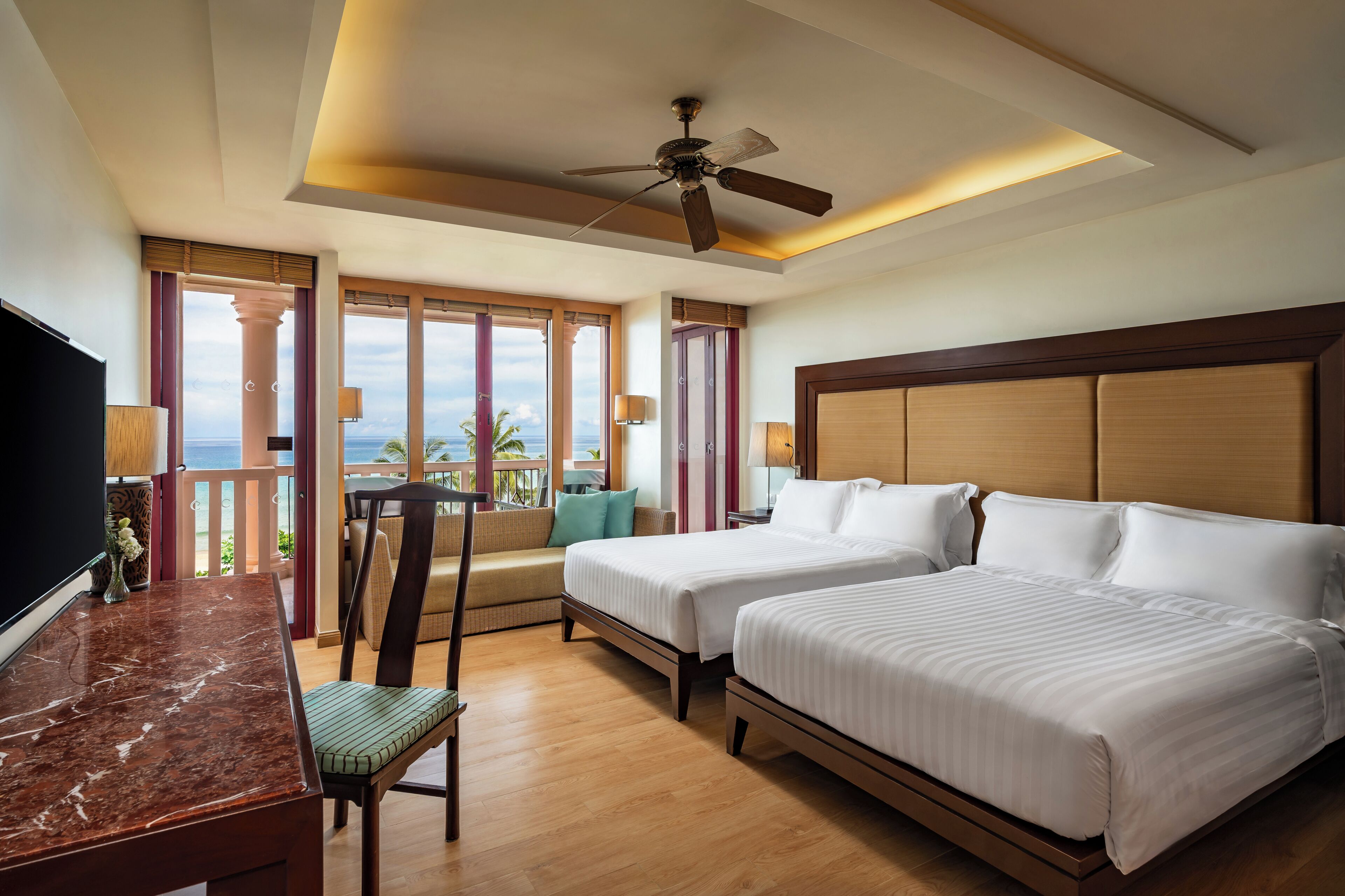 A hotel room with two beds, a desk, a chair, a TV, and a balcony with a view.