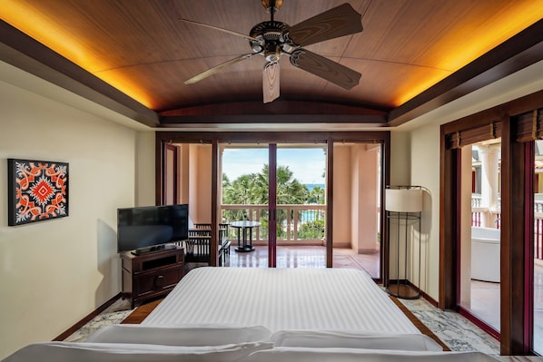 Een hotelkamer met een bed, een televisie, een balkon met uitzicht op palmbomen en een plafondventilator.