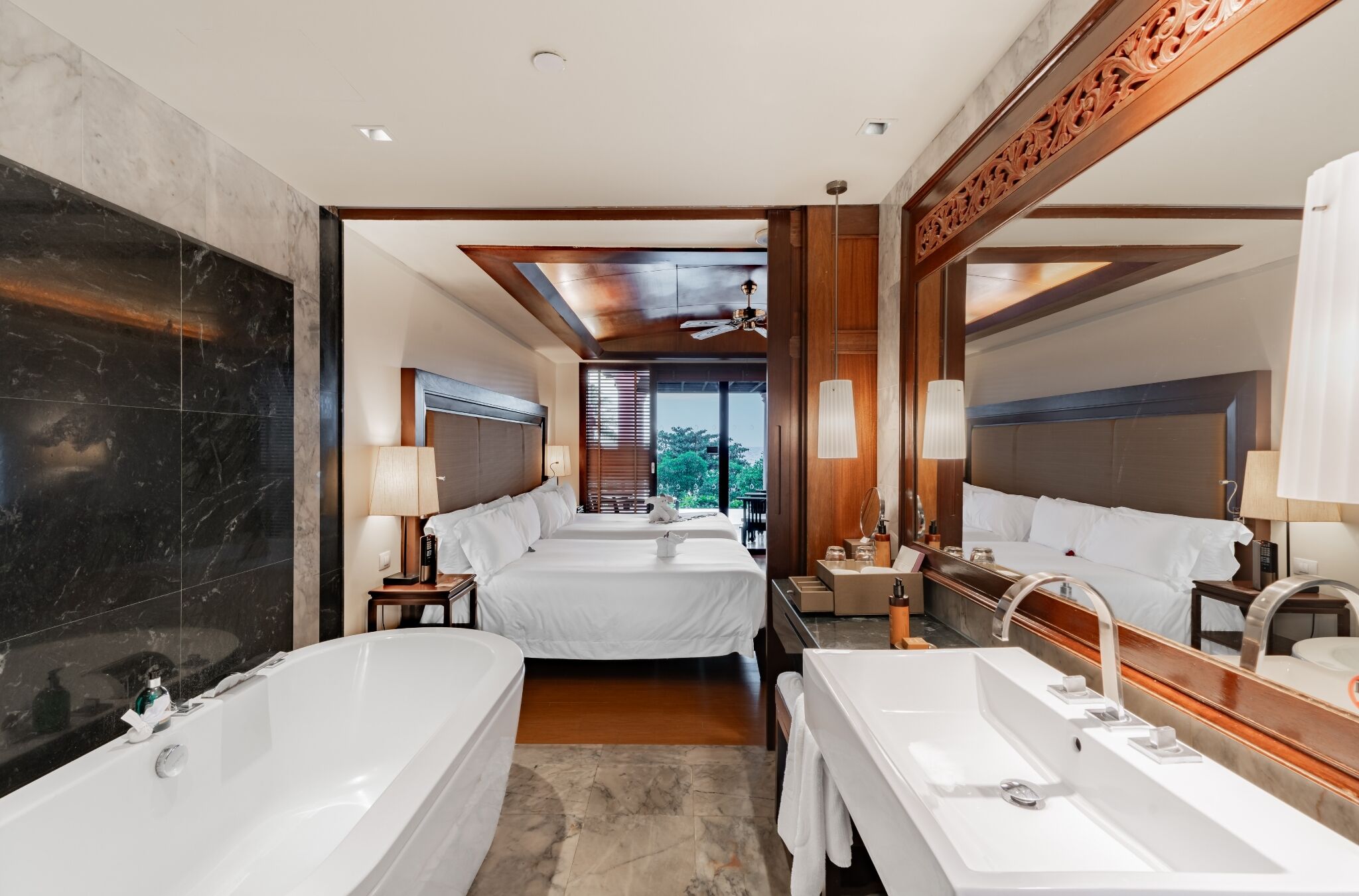 Een hotelkamer met een groot bad, twee bedden, een aparte badkamer met een spiegel en uitzicht op de omgeving.