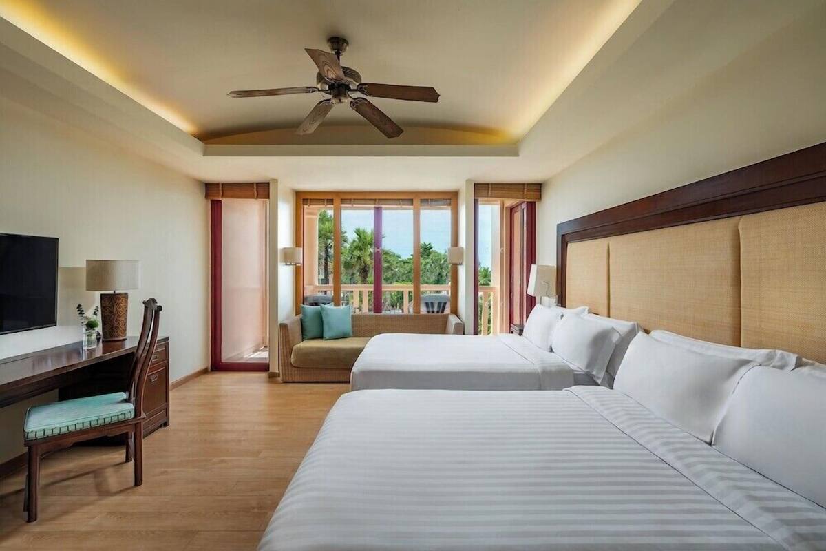 Habitación de hotel con dos camas, un escritorio con silla, televisión, ventilador de techo y ventanas amplias con vistas a zonas verdes.