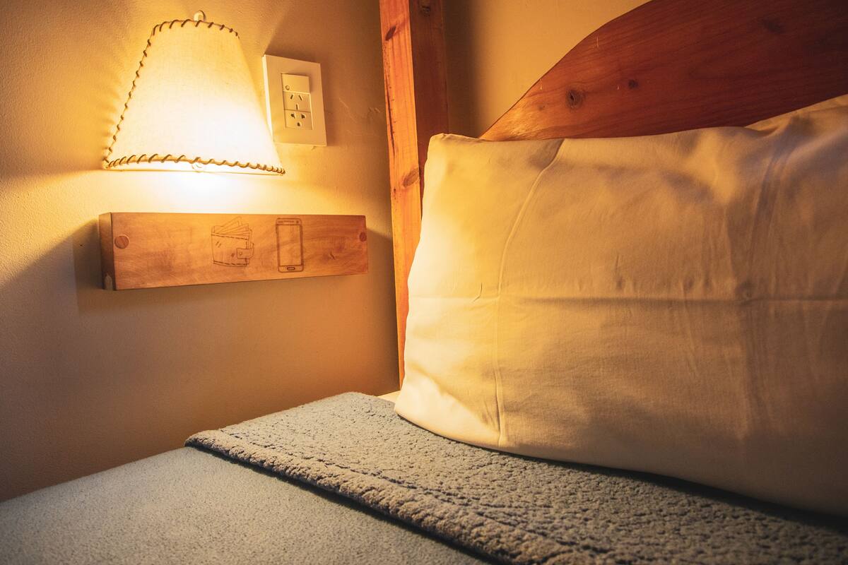 Ein ordentlich bezogenes Bett mit einem Holzkopfende, einer Nachttischlampe und einer Steckdose an der Wand.