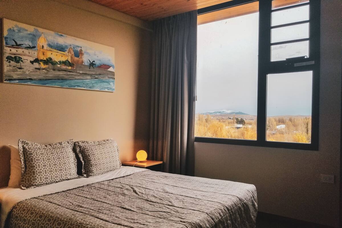 Ein Schlafzimmer mit Bett, einem Bild an der Wand, einem Nachttisch mit Lampe und einem Fenster mit Landschaftsblick.