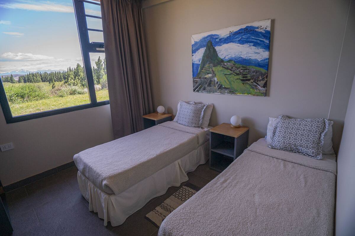 Ein Zimmer mit zwei Einzelbetten, einem Fenster mit Blick auf eine grüne Landschaft, einem Bergbild und einem kleinen Tisch mit Lampe.