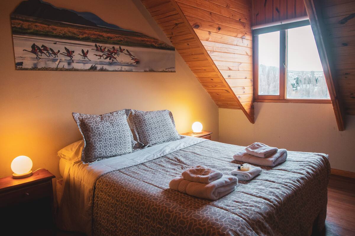 Ein Schlafzimmer mit schrägen Wänden, einem Bett mit gemustertem Überwurf, zwei Kissen, einem Nachttisch mit Lampe und einem Bild an der Wand.