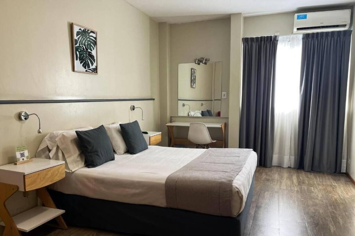 A hotel room with a bed, bedside tables, a desk, a chair, a mirror, and a window with curtains.