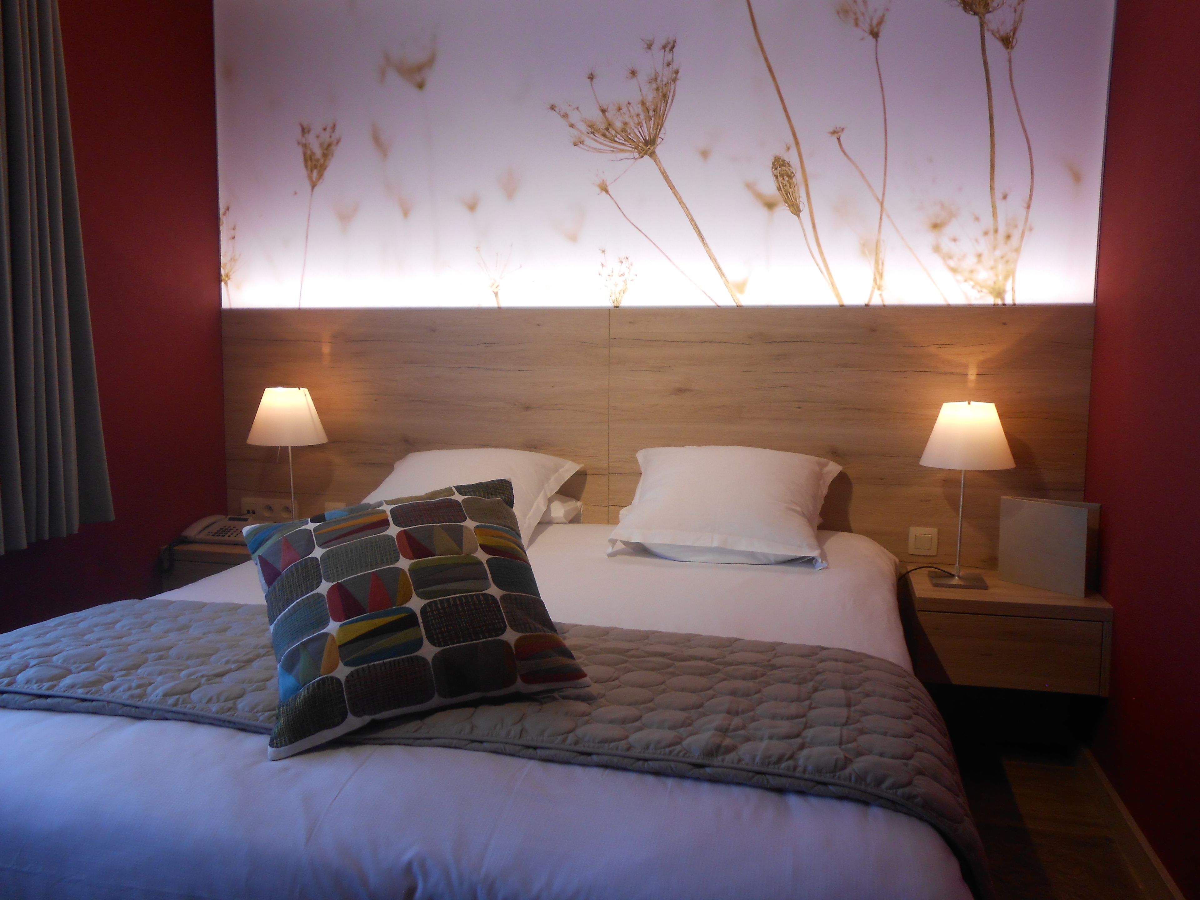 Un lit bien fait, agrémenté d’un coussin coloré, de deux lampes de chevet et d’un mur orné d’un motif floral.