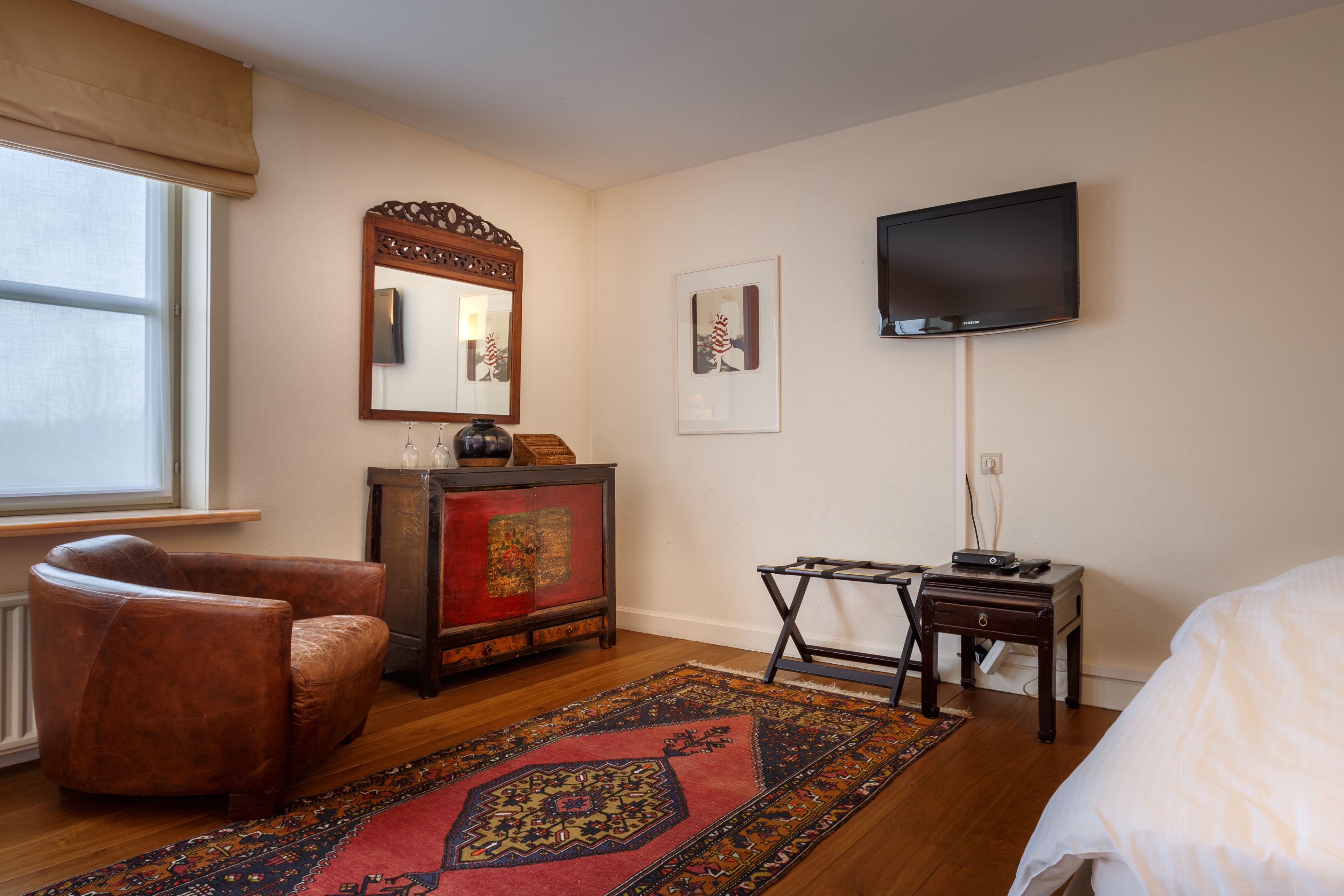 A hotel room with a wooden floor, a brown leather armchair, a red and black patterned rug, a wooden cabinet with a mirror, a flat-screen TV mounted on the wall, and a bed with white bedding.