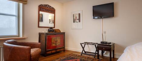 A hotel room with a wooden floor, a brown leather armchair, a red and black patterned rug, a wooden cabinet with a mirror, a flat-screen TV mounted on the wall, and a bed with white bedding.