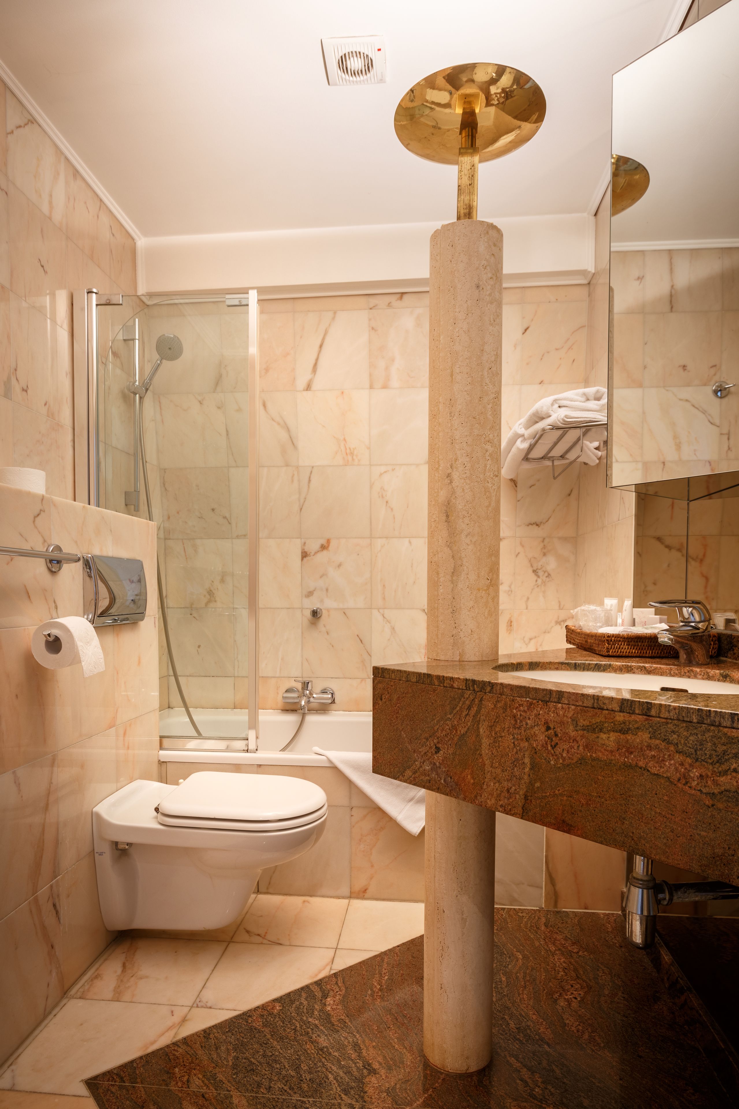 Un baño moderno con cabina de ducha de vidrio, pared con azulejos de mármol, una columna central, lavamanos con encimera de mármol y un inodoro.