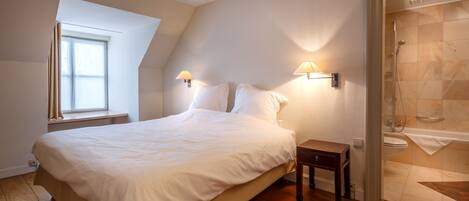 A bedroom with a large bed, bedside lamps, a small table, a window, and a shower area.