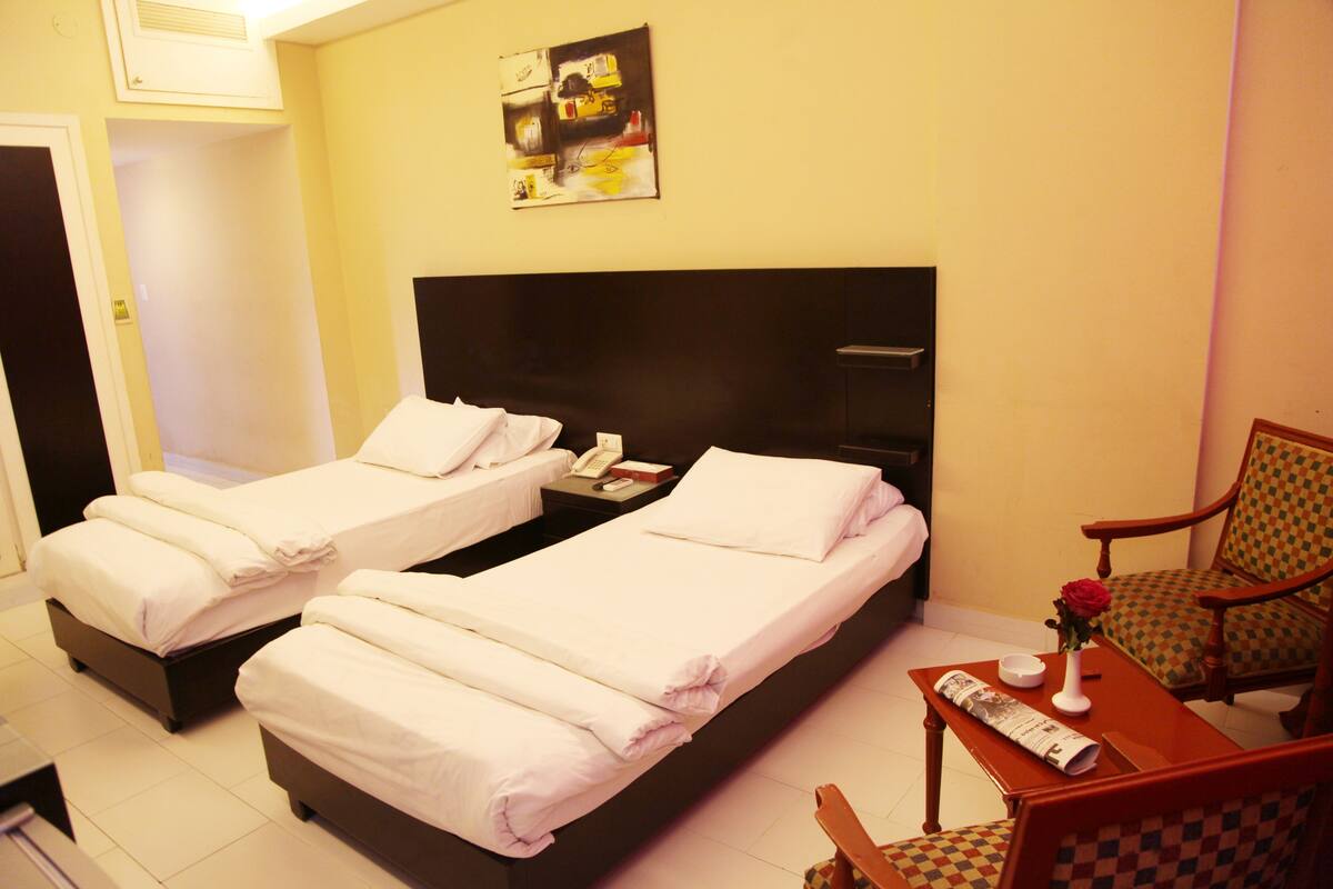 A hotel room with two single beds, a wooden chair, a small table with a vase of flowers, and a wall-mounted artwork.