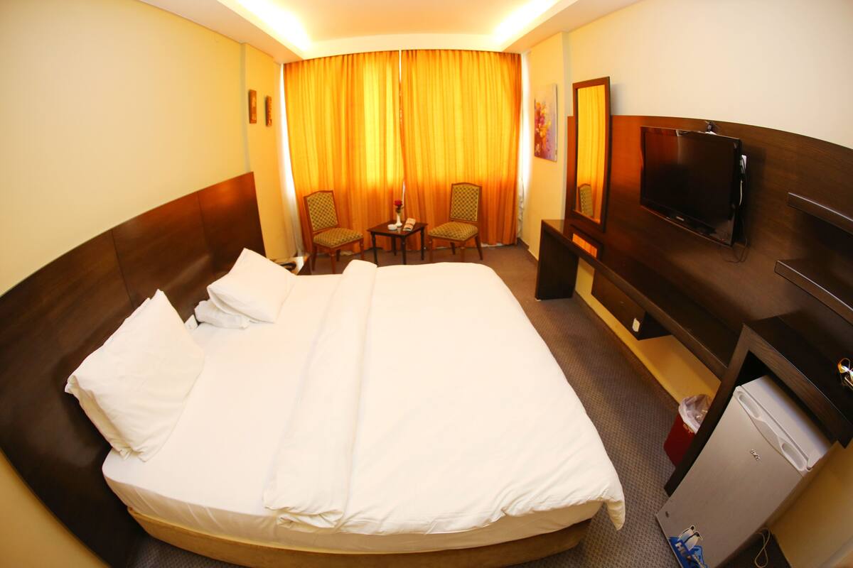 A hotel room with a large bed, two chairs, a small table, a TV, and a minibar.
