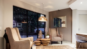 A modern living room with two patterned armchairs, a clear plastic chair, a wooden side table with a decorative tree, and a hanging pendant light. - The Lakes Boutique Lodge (Pretoria)