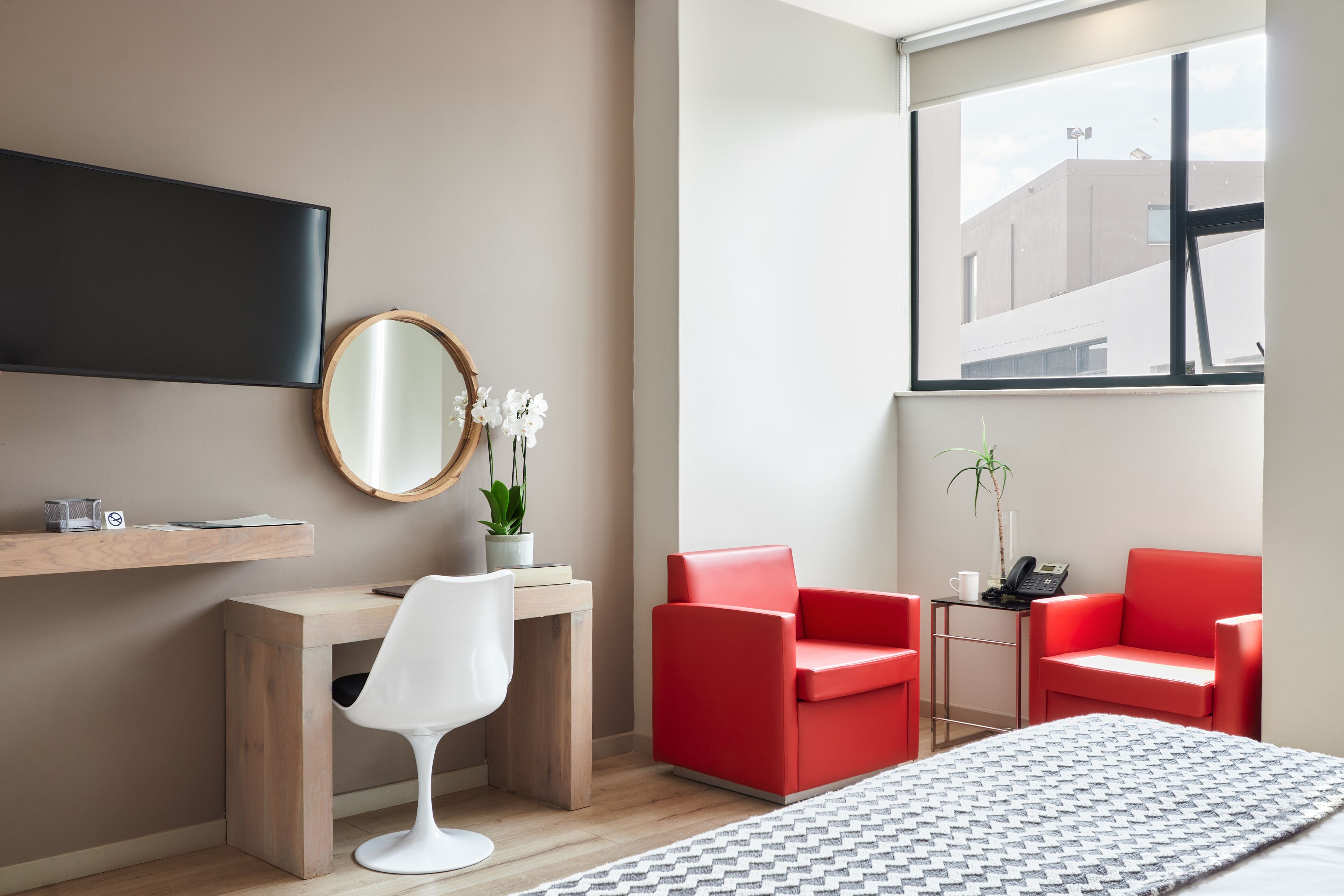 A modern hotel room with a bed, a desk, a chair, a red sofa, a TV, a mirror, and a window with a view of buildings.