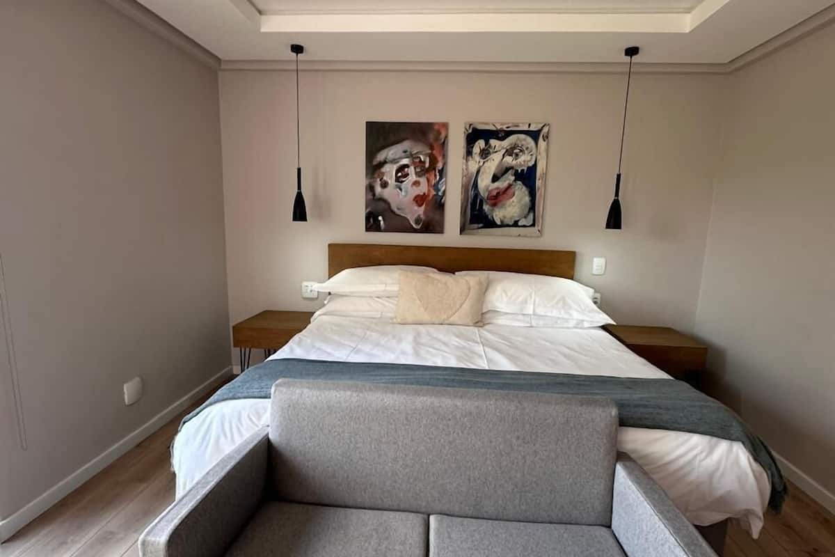 A modern bedroom with a large bed, a grey sofa, and two paintings on the wall.