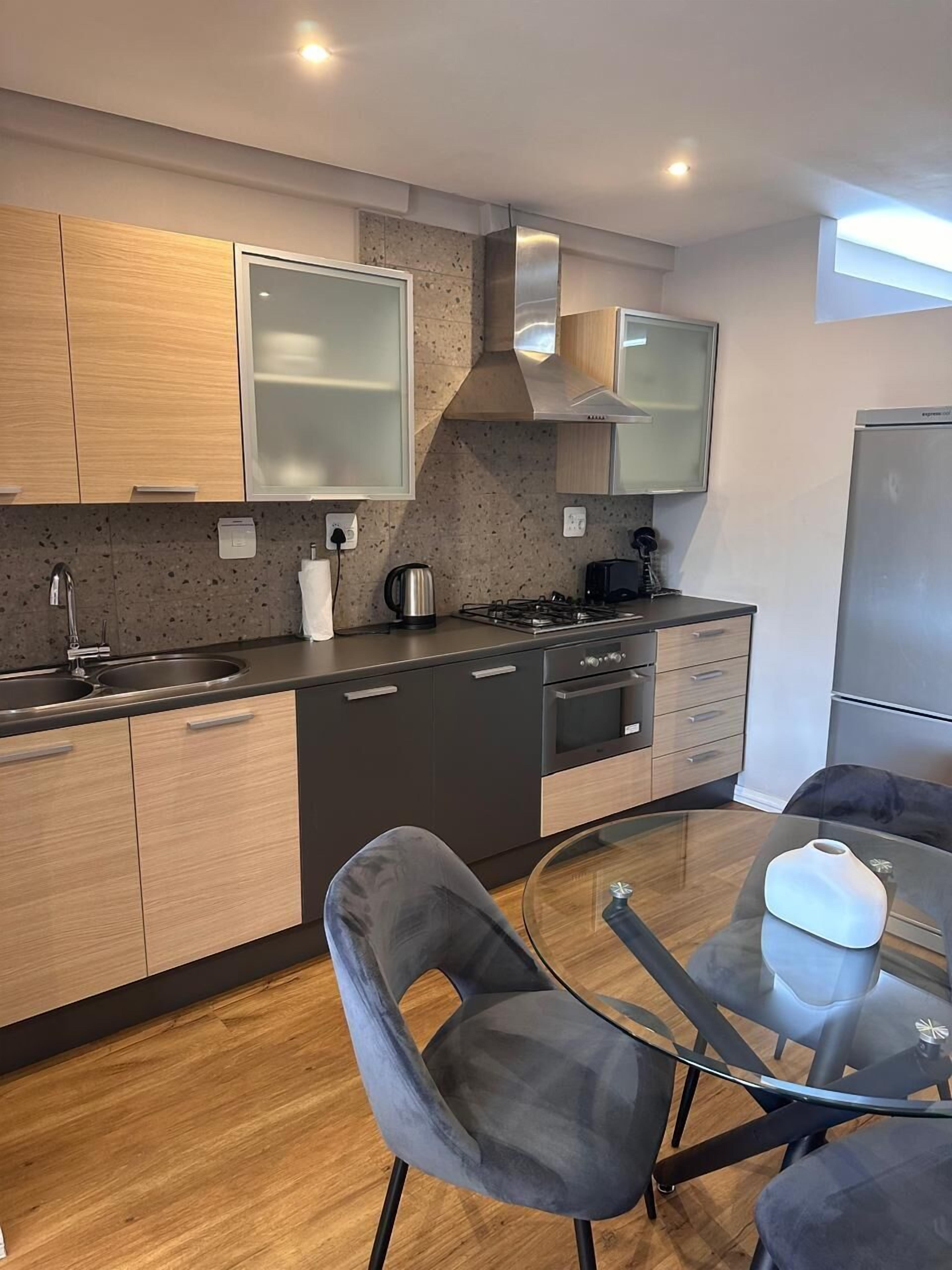 Een moderne keuken met houten kastjes, een gasfornuis, een waterkoker en een glazen eettafel met twee stoelen.