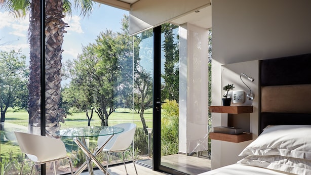 A modern bedroom with a large glass door leading to a balcony, a bed with white bedding, and a glass table with chairs. - The Lakes Boutique Lodge (Pretoria)