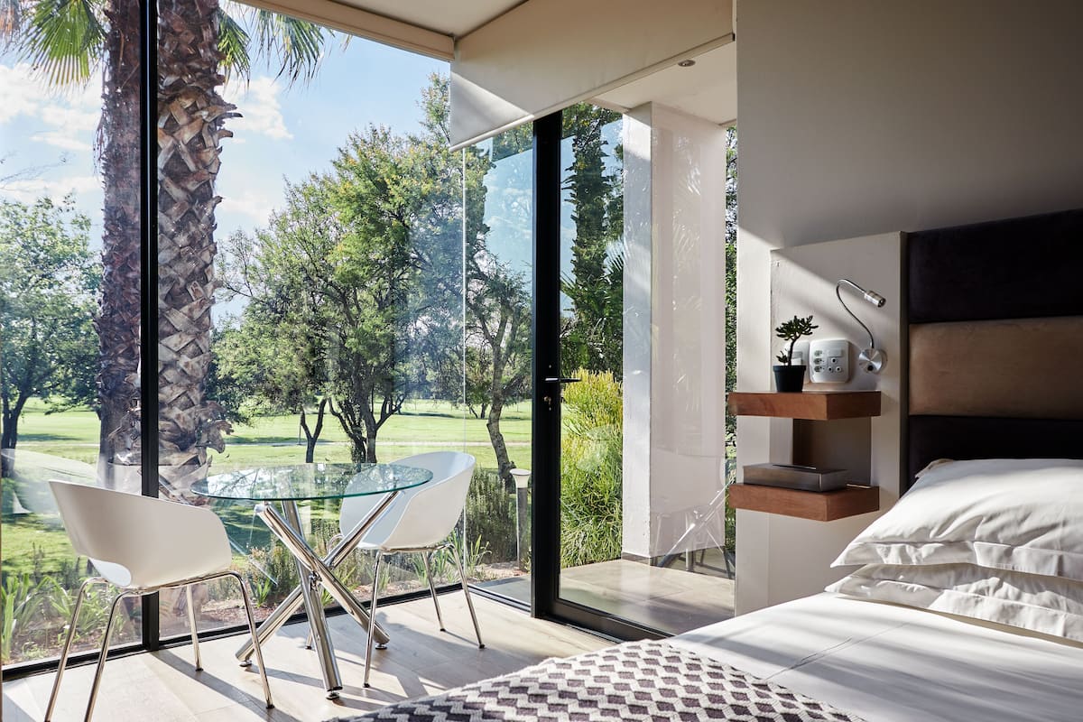 A modern bedroom with a large glass door leading to a balcony, a bed with white bedding, and a glass table with chairs.