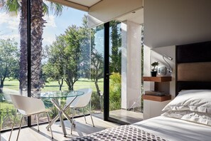 A modern bedroom with a large glass door leading to a balcony, a bed with white bedding, and a glass table with chairs. - The Lakes Boutique Lodge (Pretoria)