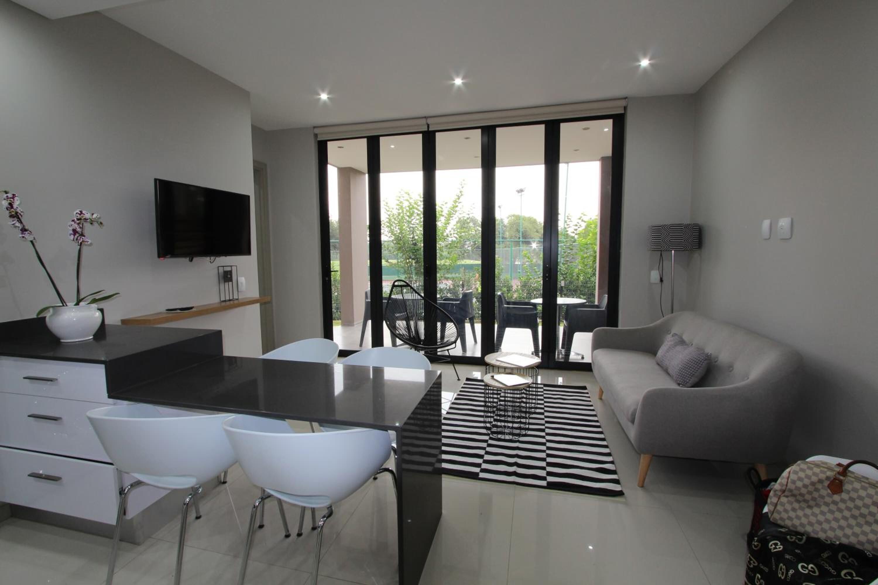 A modern living room with a grey sofa, a dining area with a black table and white chairs, and a TV mounted on the wall.