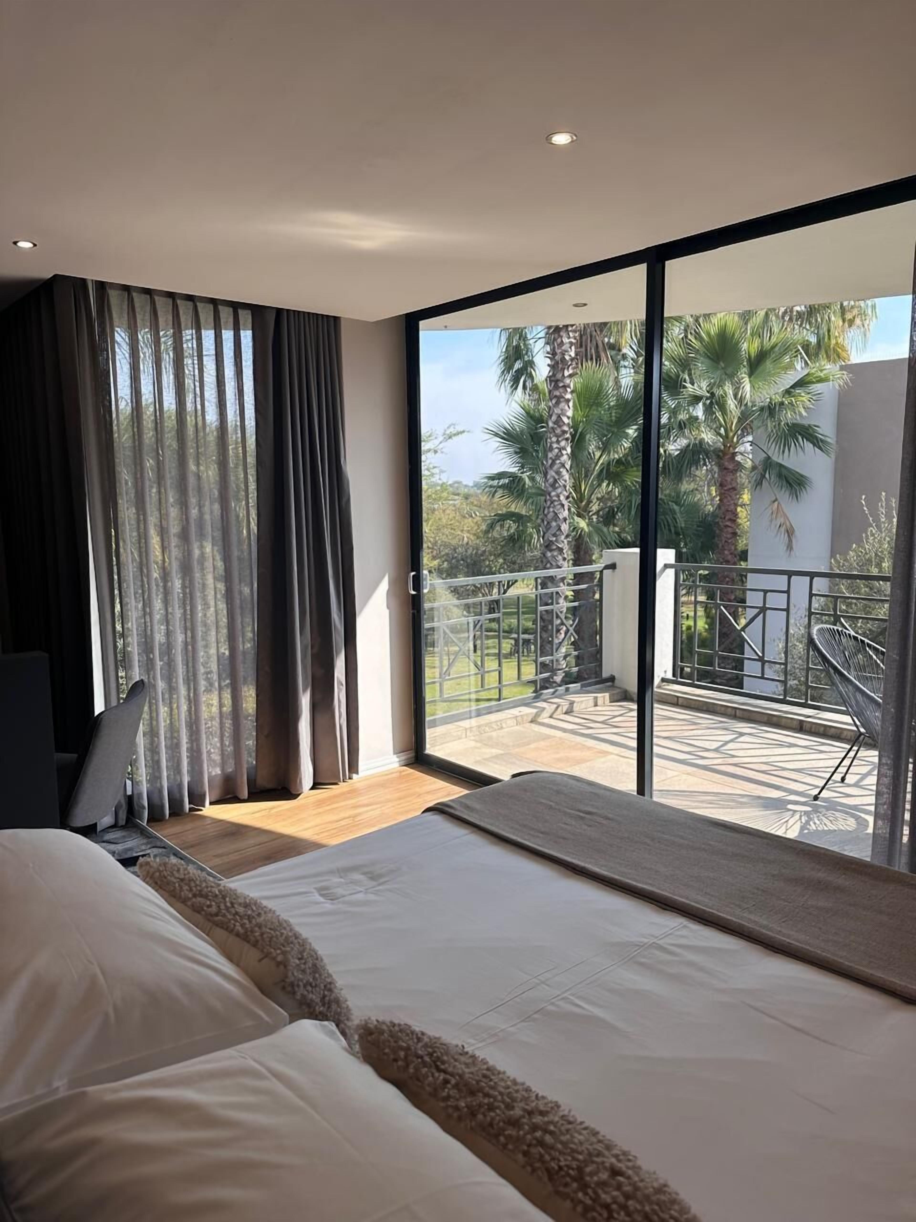 A bedroom with a large bed, a desk, and a view of a balcony with palm trees.