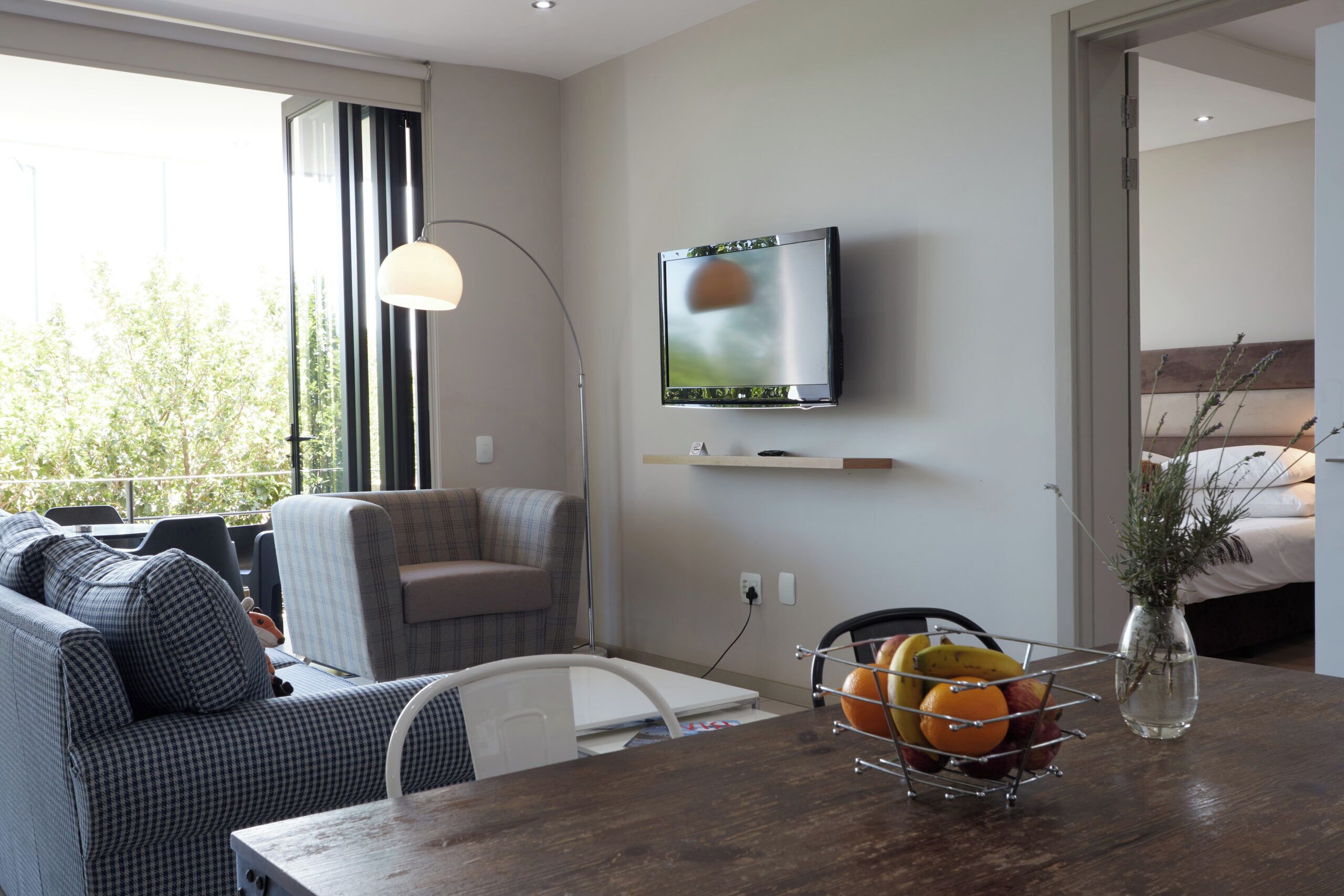 Un salon moderne avec un canapé, une table à manger ornée de fruits, un téléviseur et une vue sur l’extérieur.