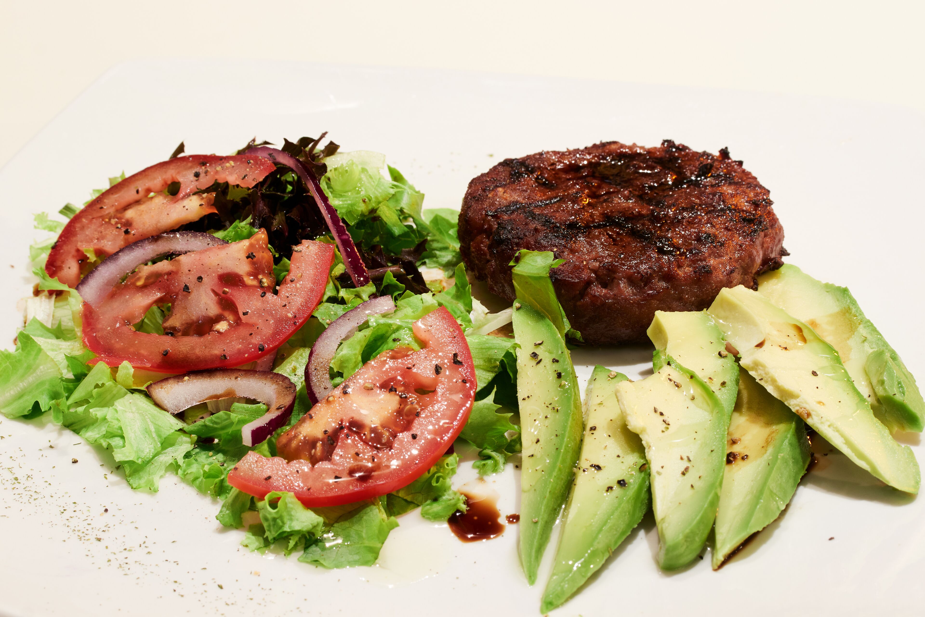Un plato con una hamburguesa a la parrilla, rebanadas de aguacate y una ensalada de lechugas mixtas con tomates y cebolla.