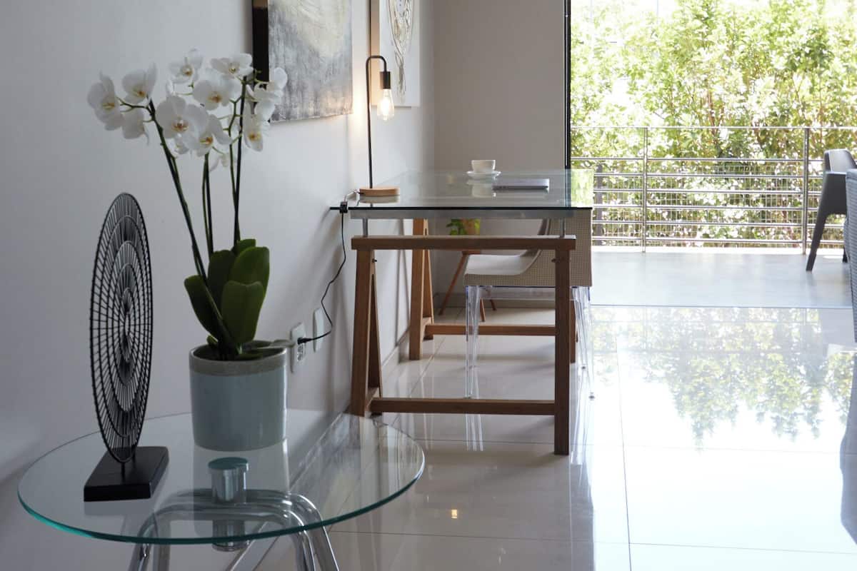 A modern living room with a glass table, a potted orchid, and a fan. There is a dining area with a wooden table and chairs, and a balcony with a view of trees.