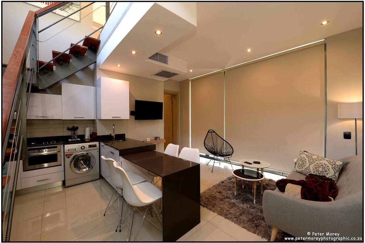 A modern kitchen with stainless steel appliances, a dining area with white chairs, and a sofa with a throw blanket.