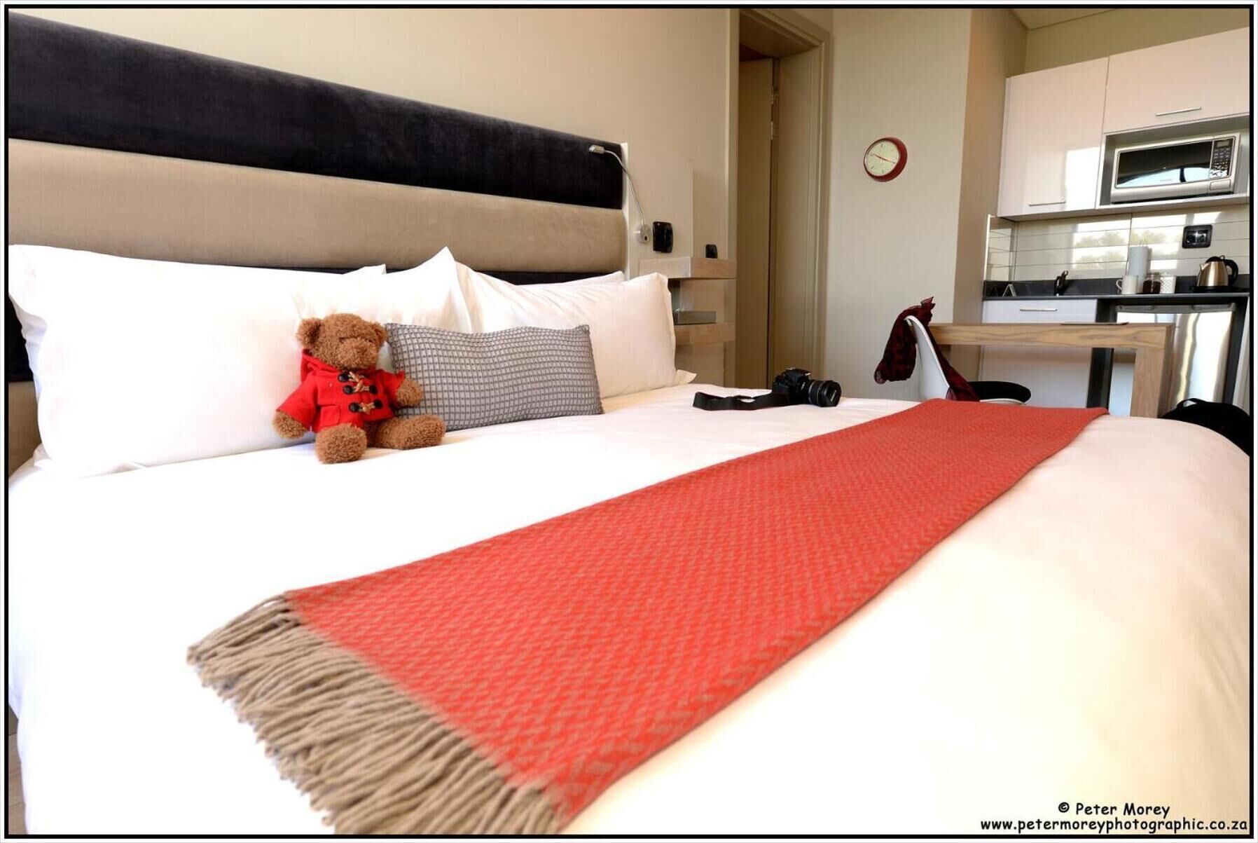 A hotel room with a bed, a teddy bear, a red blanket, a grey pillow, and a kitchen area with a microwave.