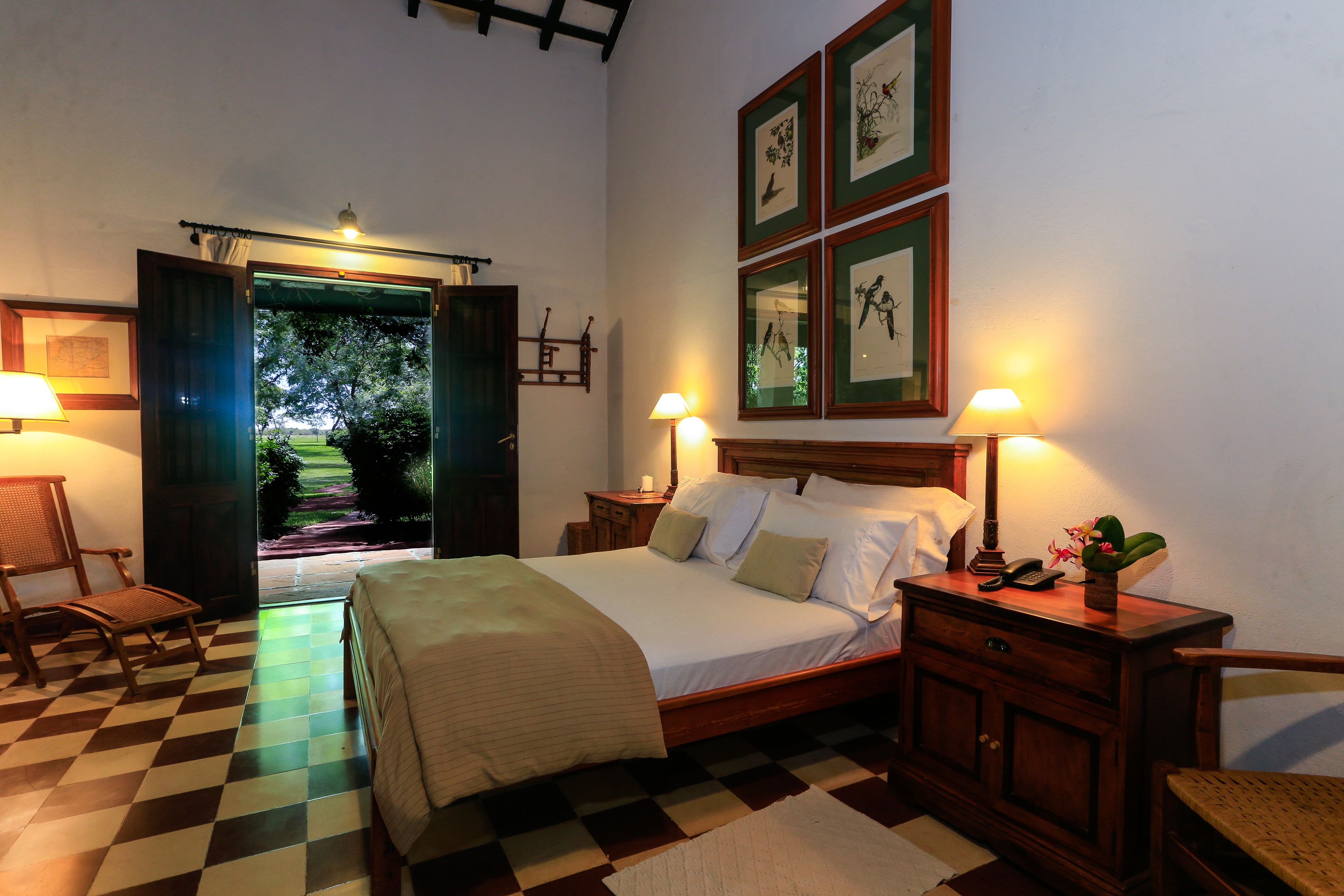A bedroom with a checkered floor, a bed with a wooden headboard, two bedside tables with lamps, and a door leading to a garden.